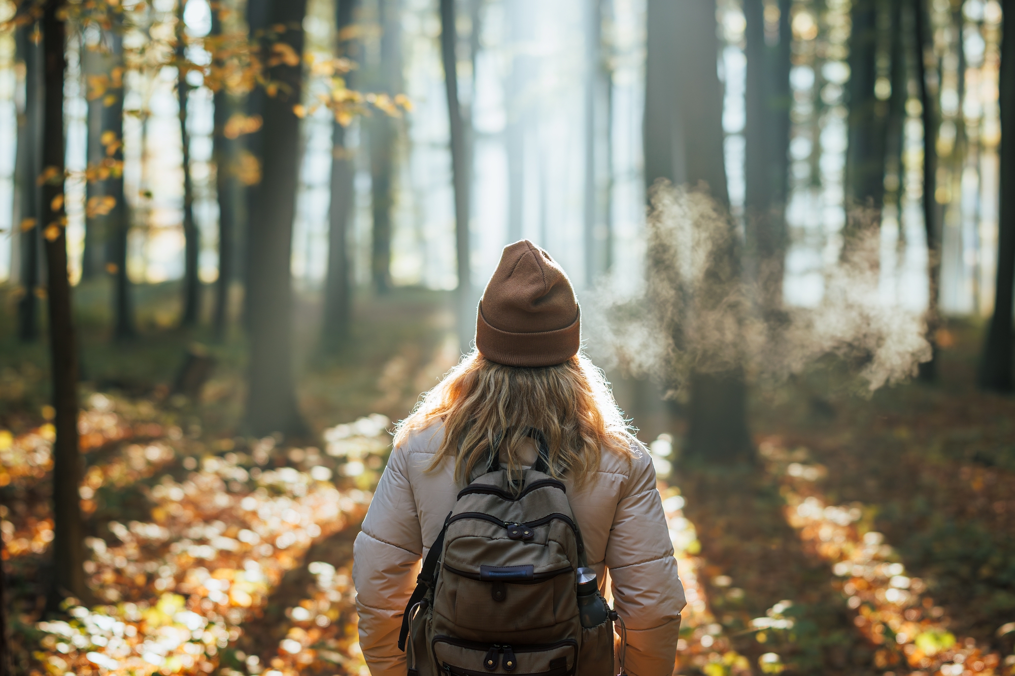 Back of woman enjoying forested hike on sunny day with backpack and beanie